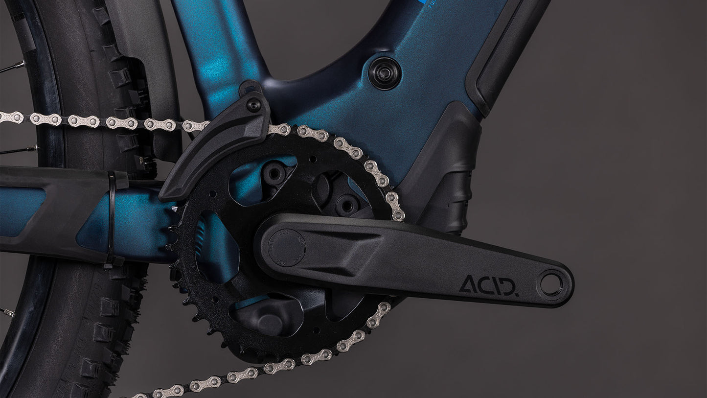 Die Nahaufnahme des CUBE Reaction Hybrid Pro 800 FE nebular'n'dazzle Trapeze 2026 Antriebs zeigt den schwarzen ACID Kurbelarm, das Kettenblatt und die Kette auf einem dunkelblauen Rahmen mit Shimano XT Schaltung vor einem schwarzen Hintergrund.