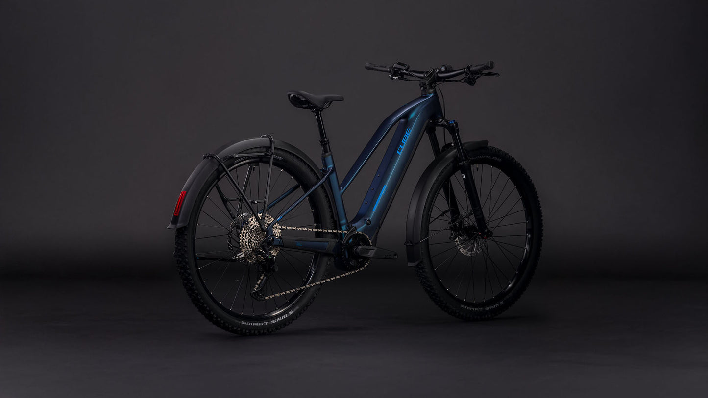 Das CUBE Reaction Hybrid Pro 800 FE nebular'n'dazzle Trapeze 2026 ist ein modernes dunkelblaues E-Bike Mountainbike mit schwarzen Schutzblechen, dicken Reifen, Shimano XT Schaltung und einer Hinterradkassette, die sich vor einem dunklen, neutralen Hintergrund präsentiert.
