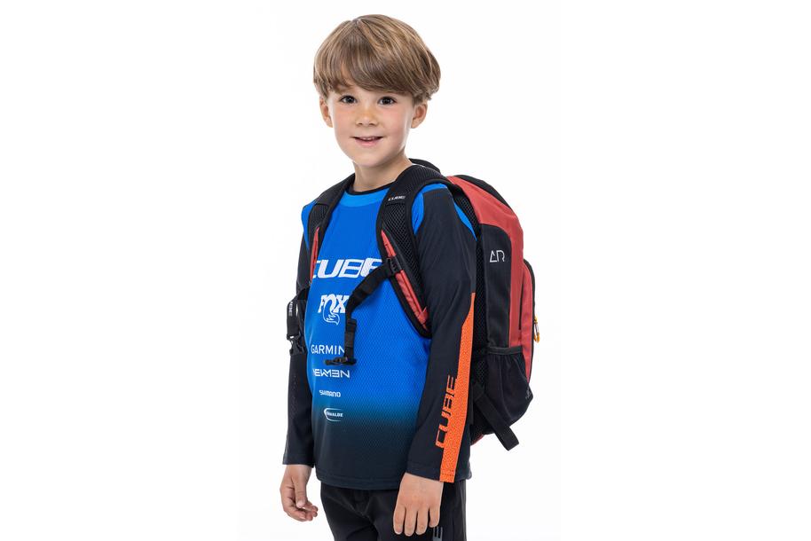 Ein Junge mit hellbraunem Haar lächelt, während er ein blau-schwarzes langärmeliges Hemd trägt und den CUBE Rucksack PURE 6 ROOKIE red'n'black von CUBE vor einem schlichten weißen Hintergrund steht.