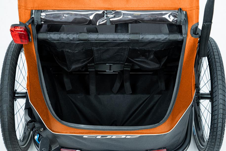 Nahaufnahme des hinteren Staufachs des CUBE Kinder Fahrradanhängers Double CMPT X Actionteam von Cube in Orange. Zu sehen sind zwei große Räder, schwarze Netztaschen im Inneren, ein rotes Rücklicht, Insektenschutz und der Cube Schriftzug am Boden.