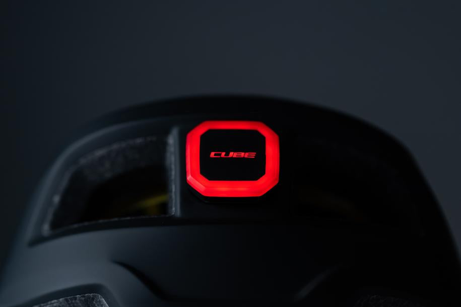 Nahaufnahme des CUBE Helm Rücklicht CMPT von Cube: ein schwarzer Helm mit rot leuchtendem LED-Rücklicht, CUBE-Branding und dunkler Kulisse - ideal für Radsportfans in Göttingen oder im CUBE Store vor Ort.
