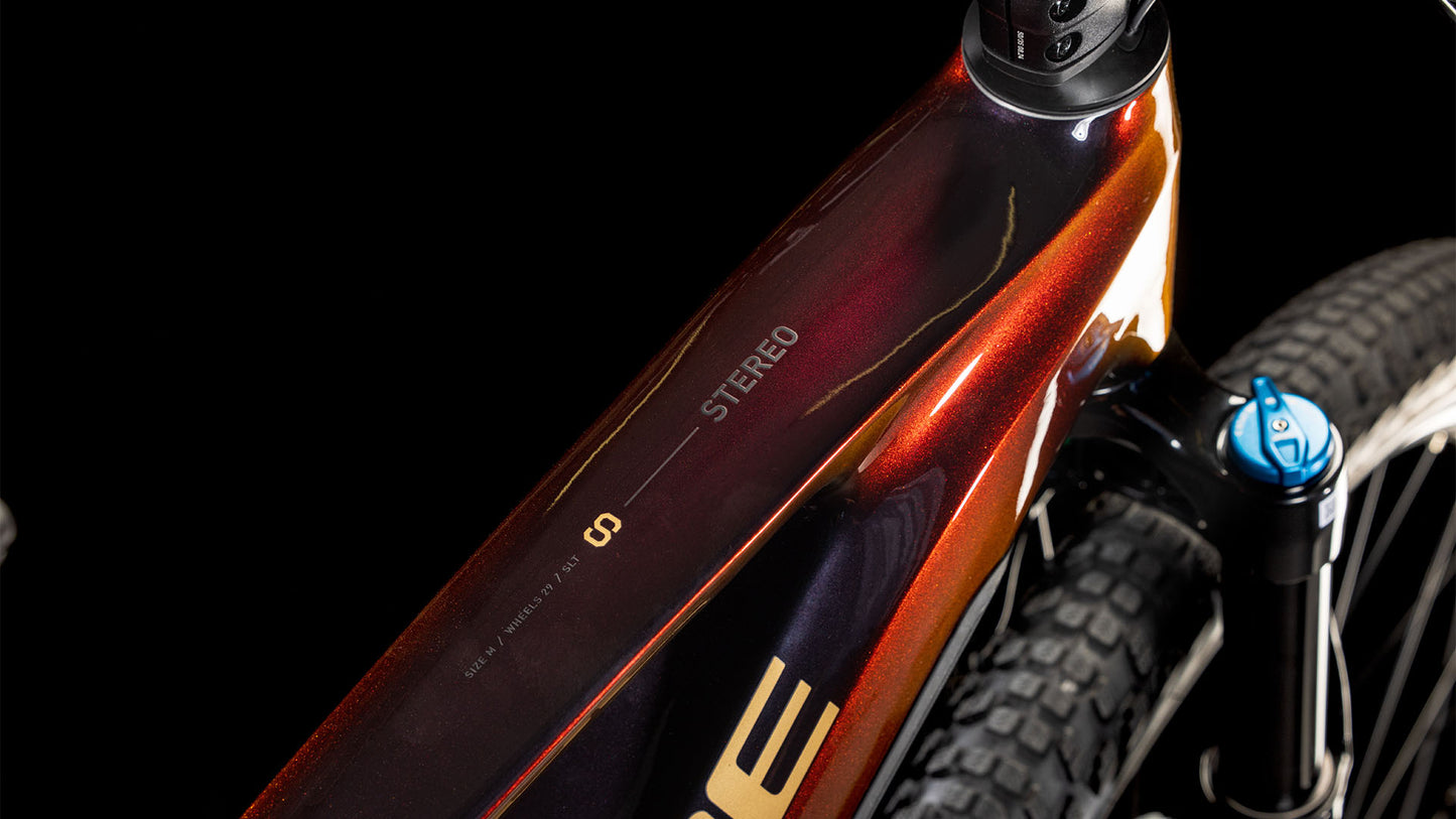 Die Nahaufnahme des CUBE Stereo Hybrid ONE22 SLT 800 solareclipse'n'gold 2025 zeigt das rot-orange glänzende Oberrohr mit "STEREO"-Schriftzug sowie einen Blick auf die Federgabel und den reifenbetonten Bosch CX-Antrieb für E-Mountainbiker.