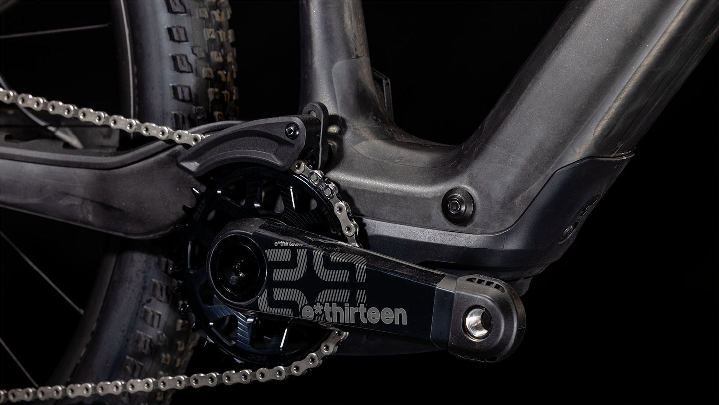 Nahaufnahme einer CUBE AMS Hybrid ONE44 C:68X SLT 400X 29-Kurbelgarnitur und eines e*thirteen-Kettenblatts am Carbon'n'Golddust-Rahmen, die einen Teil des Hinterradreifens und ein superleichtes MTB-Fully vor schwarzem Hintergrund zeigt.