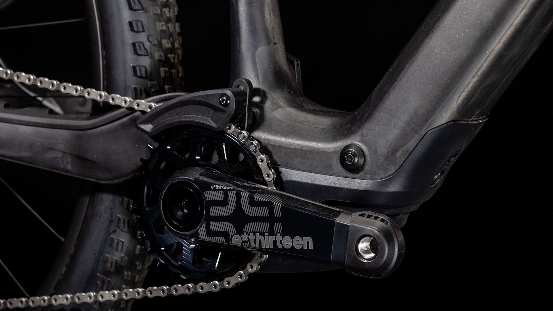 Nahaufnahme einer CUBE AMS Hybrid ONE44 C:68X SLT 400X 29-Kurbelgarnitur und eines e*thirteen-Kettenblatts am Carbon'n'Golddust-Rahmen, die einen Teil des Hinterradreifens und ein superleichtes MTB-Fully vor schwarzem Hintergrund zeigt.