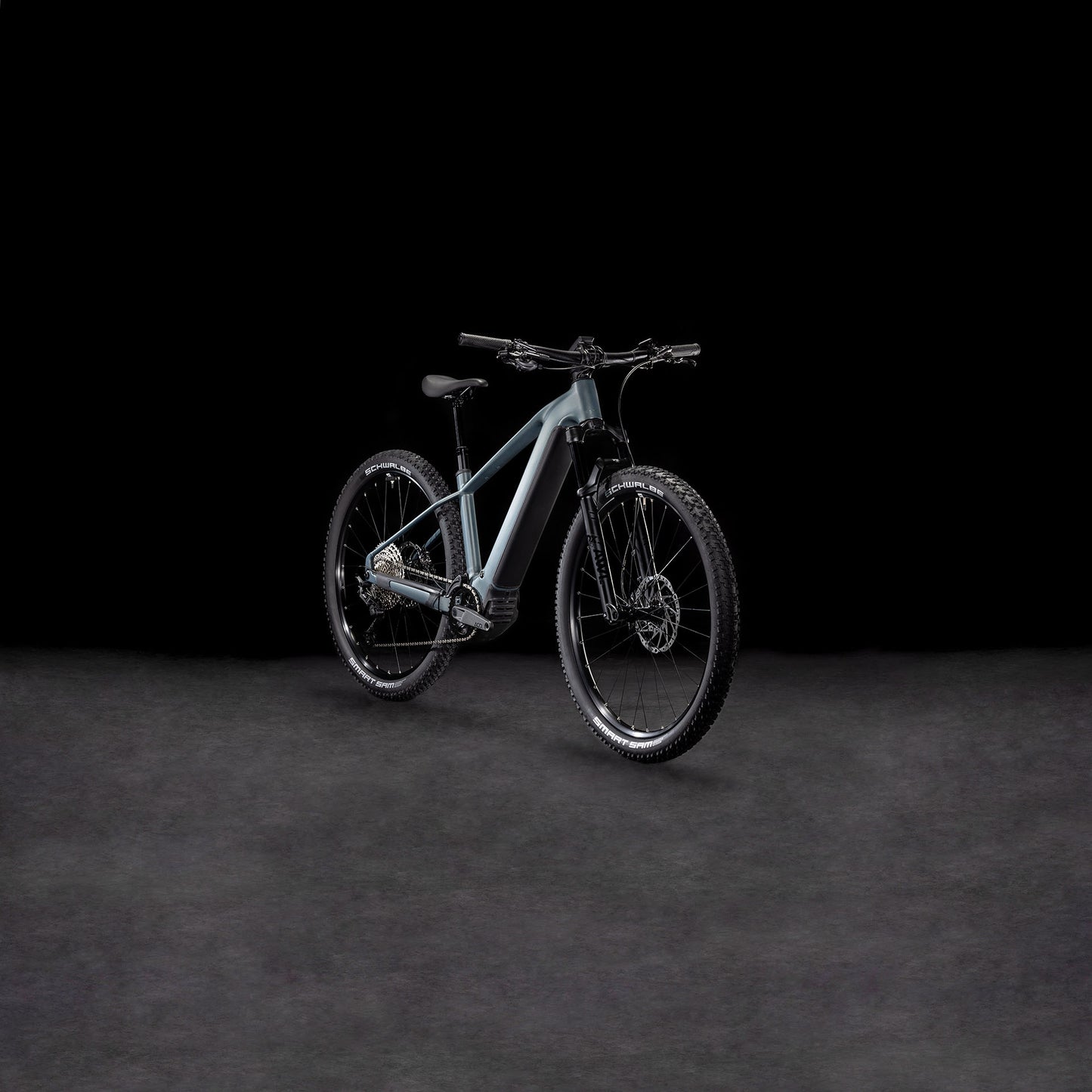 Das CUBE Reaction Hybrid SLX 800 sagebrushgreen'n'prism 2025 E-Hardtail Mountainbike mit einem 800 Wh PowerTube Akku steht auf einer dunklen, strukturierten Oberfläche vor schwarzem Hintergrund.