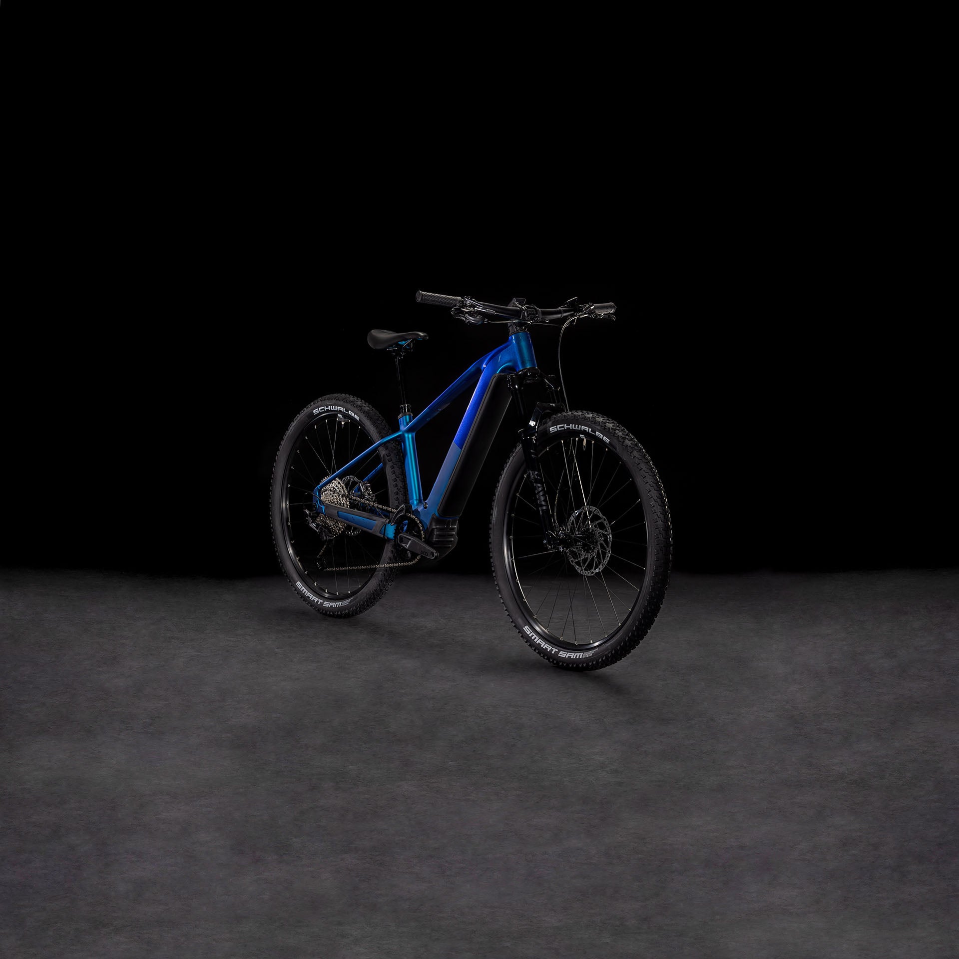 Das CUBE Reaction Hybrid SLX 800 nebula'n'blue 2025 Mountainbike mit dicken Reifen und einem leistungsstarken Bosch CX Motor steht auf einer dunkel strukturierten Oberfläche vor einem schwarzen Hintergrund.
