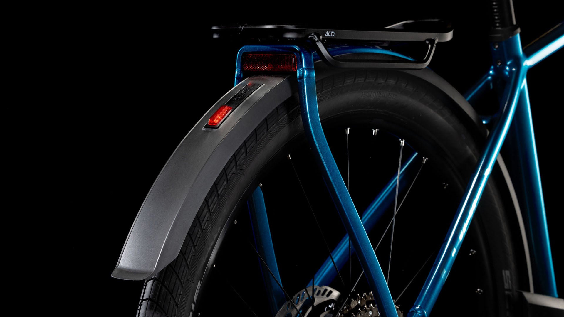 Nahaufnahme des Hinterrads und Schutzblechs des CUBE Kathmandu Hybrid EX 800 electricblue´n´chrome 2025. Zu sehen sind der blaue Rahmen, der schwarze Reifen, das silberne Schutzblech und der rote Rückstrahler vor einem schwarzen Hintergrund.