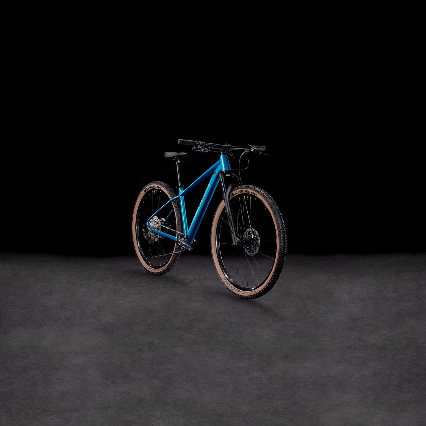 Das CUBE Reaction Pro metallicteal'n'holo 2025 Mountainbike mit hellbraunen Reifen, hydraulischen Shimano-Scheibenbremsen und einer RockShox Judy Silver Luftgabel wird auf einer dunkel strukturierten Oberfläche vor einem schwarzen Hintergrund dargestellt.