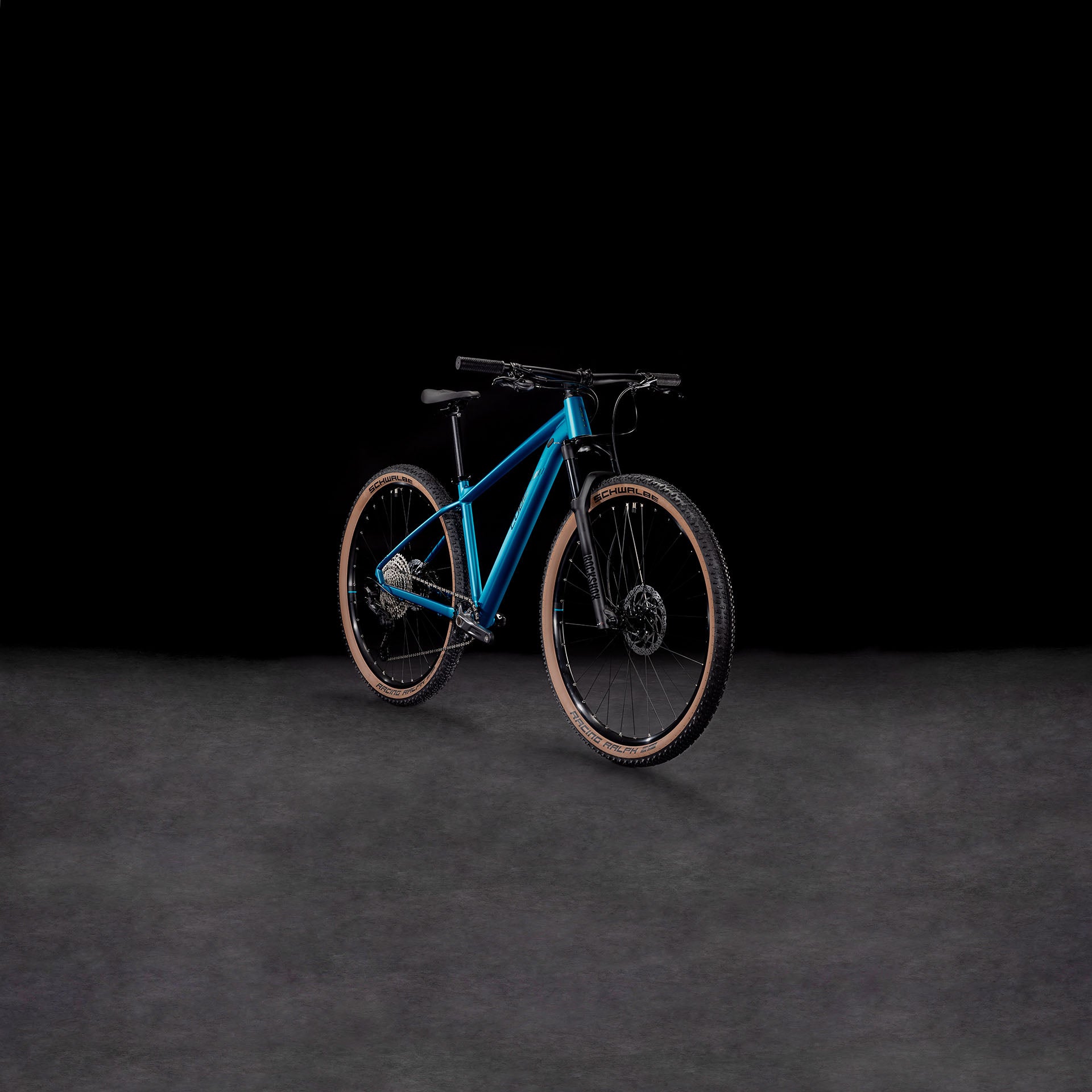 Das CUBE Reaction Pro metallicteal'n'holo 2025 Mountainbike mit hellbraunen Reifen, hydraulischen Shimano-Scheibenbremsen und einer RockShox Judy Silver Luftgabel wird auf einer dunkel strukturierten Oberfläche vor einem schwarzen Hintergrund dargestellt.