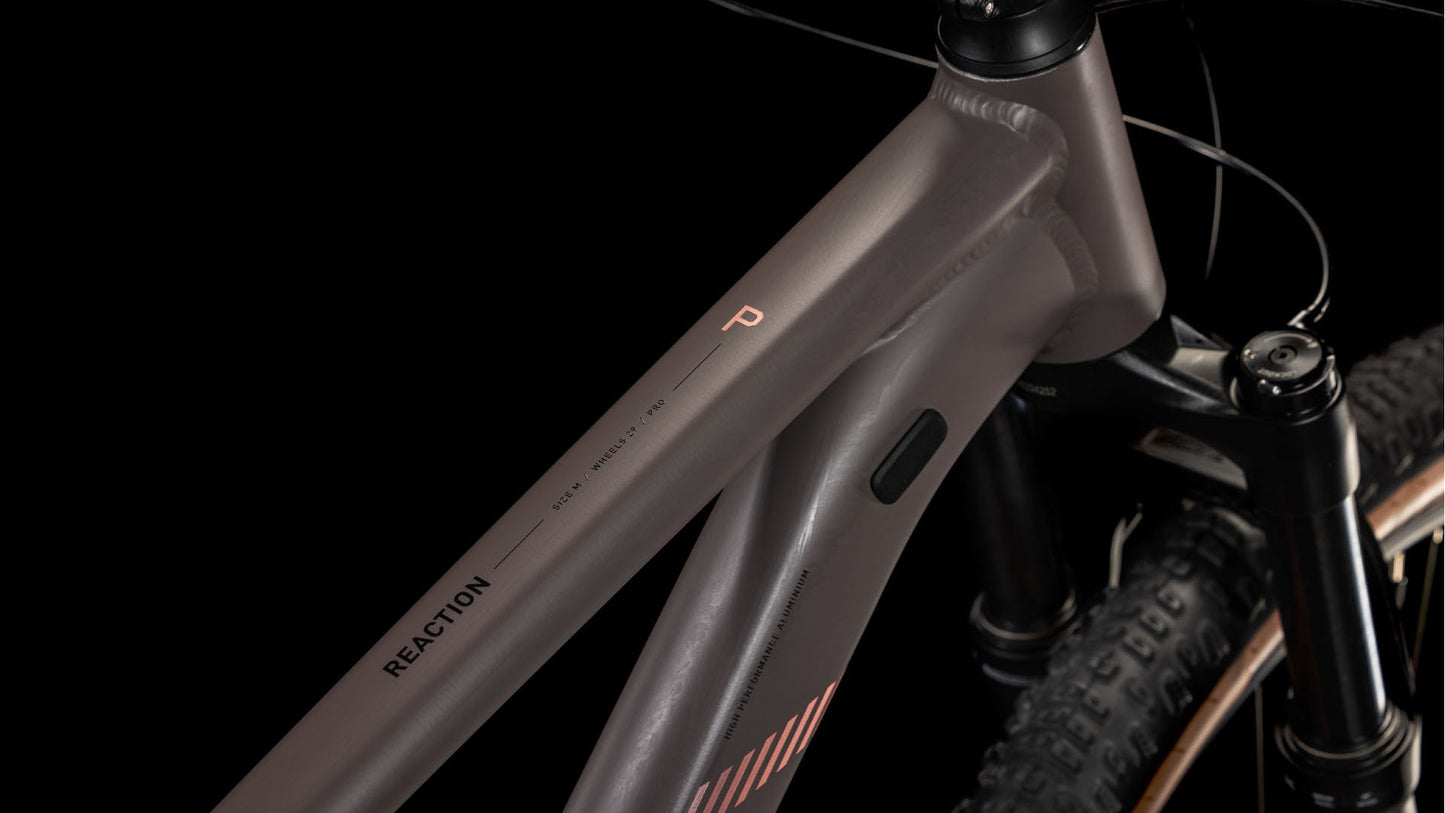 Nahaufnahme des matten cedar´n´blush CUBE Reaction Pro 2025-Rahmens mit „REACTION“ auf dem Oberrohr, der einen Teil der RockShox Judy Silver Luftfedergabel und einen grobstolligen Vorderreifen vor einem schwarzen Hintergrund zeigt.
