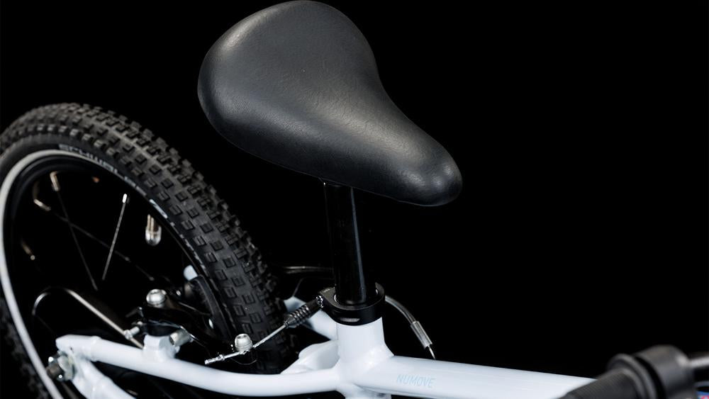 Nahaufnahme eines schwarzen Fahrradsitzes und eines Teils des Hinterrads vor einem schwarzen Hintergrund. Der weiße Rahmen des CUBE Numove 140 flashwhite'n'pink 2025 sticht hervor, und der Reifen von CUBE hat ein Noppenprofil.