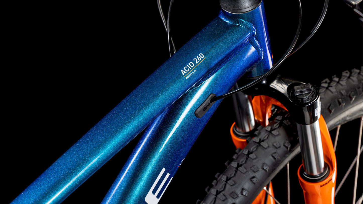 Nahaufnahme des CUBE Acid 260 Disc actionteam 2025 Mountainbike-Rahmens in Blau mit schwarzen Zügen und einer orangefarbenen Federgabel mit Noppenreifen vor einem schwarzen Hintergrund.