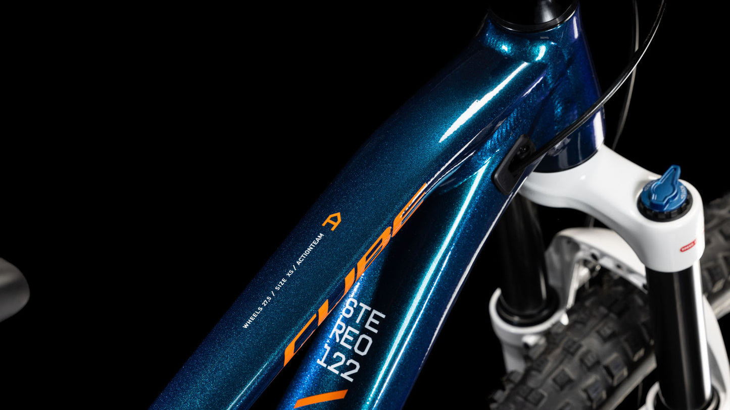 Nahaufnahme eines blaumetallicfarbenen CUBE Stereo ONE22 Rookie actionteam 2025 Mountainbike-Rahmens mit orangefarbenem und weißem Branding, der die vordere Federgabel und die Stollenreifen zeigt - ideal für Mountainbike-Fans - vor einem schwarzen Hintergrund.
