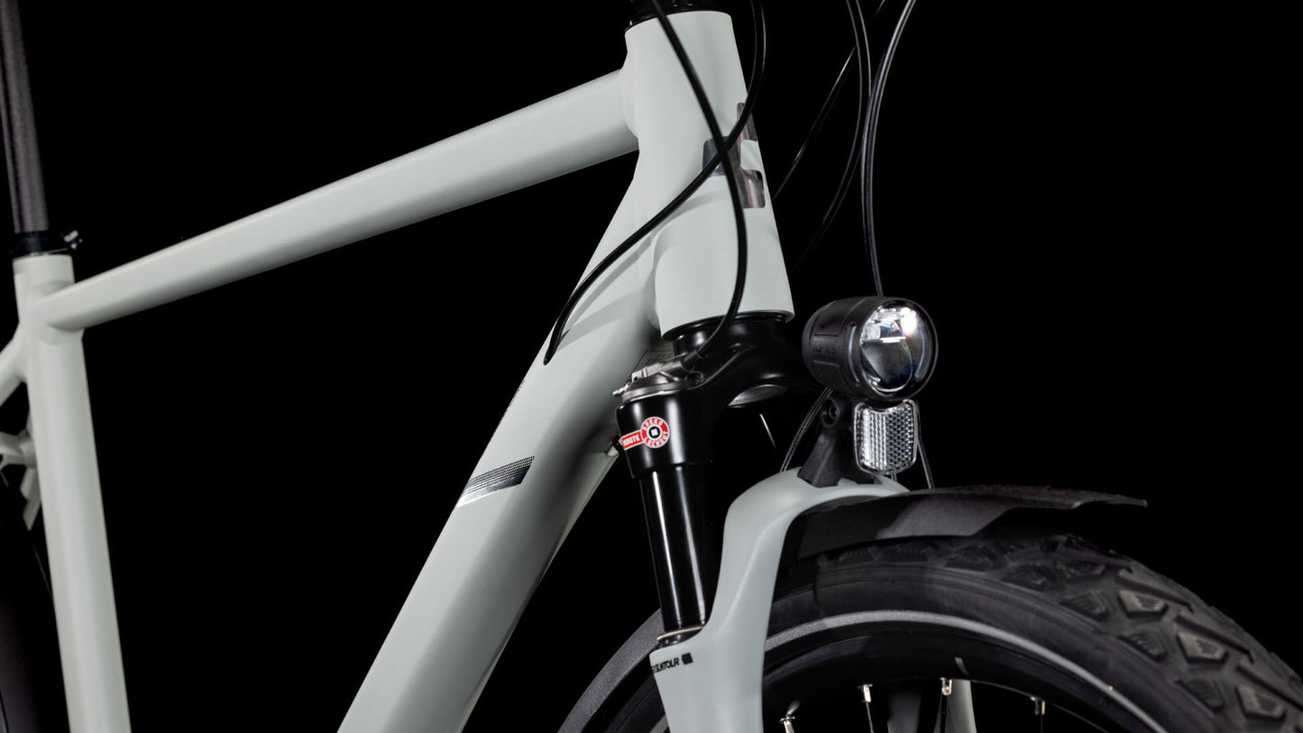 Nahaufnahme der Frontpartie des CUBE Nature EXC Allroad seasalt'n'pyrite 2025, die den hellgrauen Rahmen, die Suntour-Federgabel, den Scheinwerfer, den Reifen und die Bremszüge vor einem schwarzen Hintergrund zeigt.