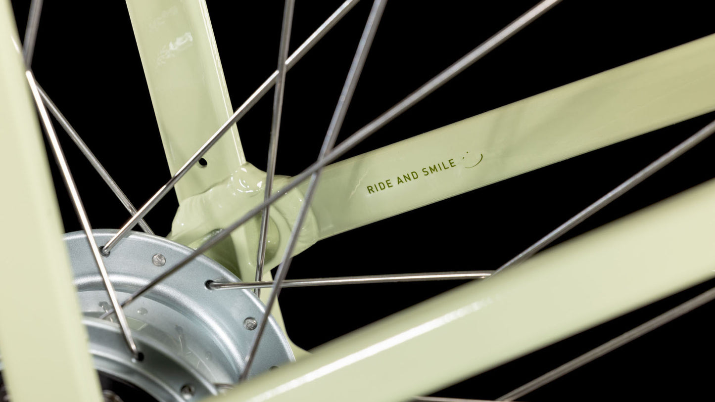 Nahaufnahme eines hellgrünen CUBE ella matchagreen'n'green Easy Entry 2025 Fahrradrades mit Metallspeichen und "RIDE AND SMILE" auf dem Rahmen. Der Hintergrund ist schwarz, und das CUBE Fahrrad hat eine 8-Gang Nabenschaltung für reibungsloses Fahren.
