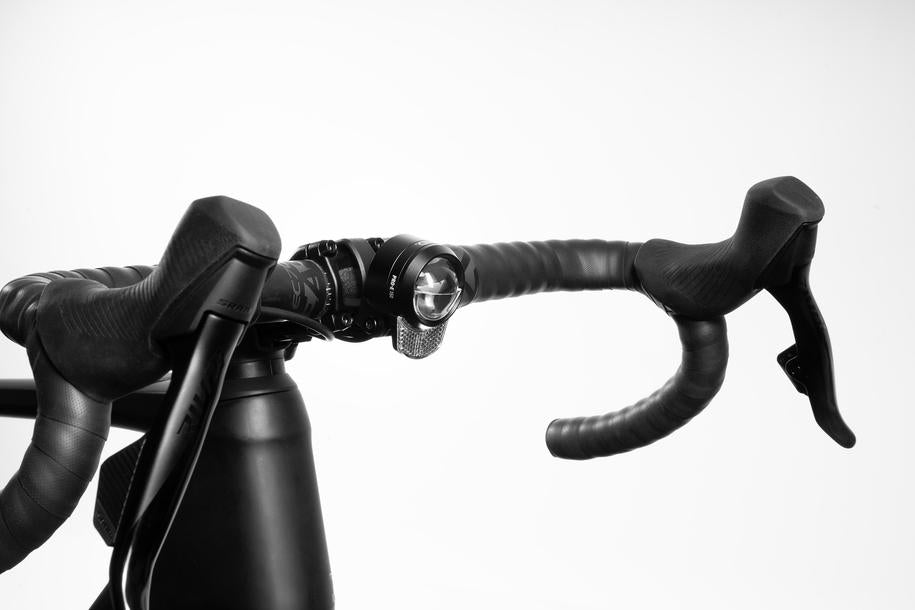 Nahaufnahme eines schwarzen Rennradlenkers mit strukturiertem Griffband, Bremshebeln und dem ACID E-Bike Frontlicht PRO-E 150 X-Connect von ACID mit hoher LED-Leistung vor einem schlichten weißen Hintergrund.