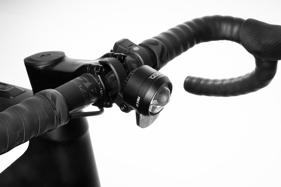 Nahaufnahme eines Fahrradlenkers mit umwickelten Griffen, integrierten Bedienelementen und einem ACID E-Bike Frontlicht PRO-E 150 X-Connect, fotografiert vor einem schlichten weißen Hintergrund.