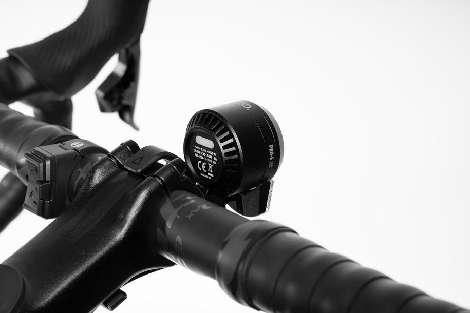 Nahaufnahme eines ACID E-Bike Frontlichts PRO-E 150 X-Connect, montiert am Fahrradlenker in Vorbaunähe, mit starker LED-Leistung. Der Hintergrund ist weiß und leicht verschwommen.