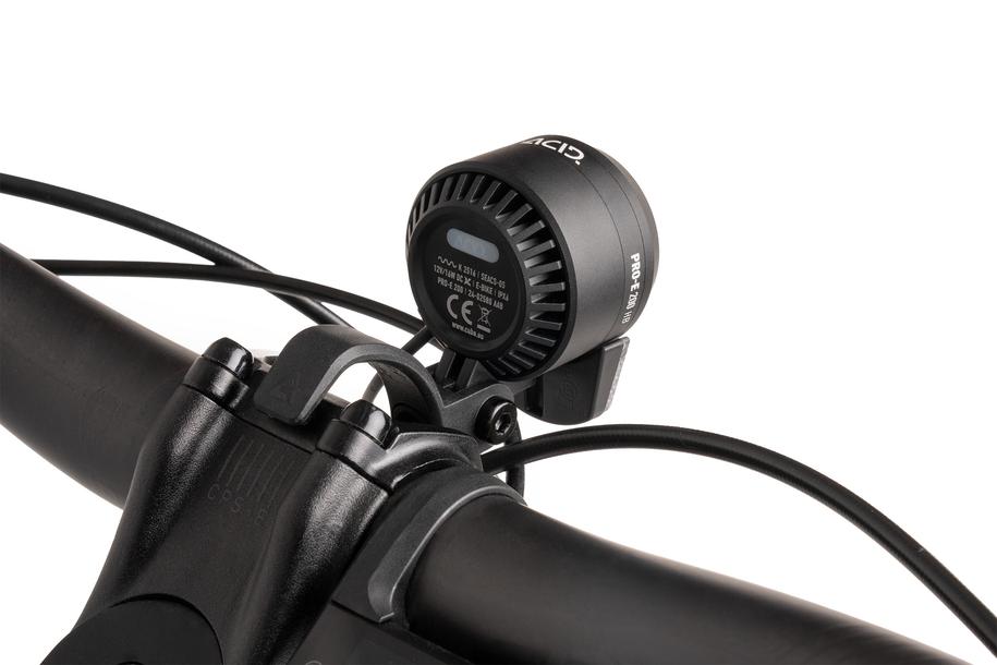 Nahaufnahme eines schwarzen Lenkers mit dem ACID E-Bike Frontlicht PRO-E 200 High Beam X-Connect von ACID. Das Frontlicht zeichnet sich durch ein rundes Design, eine belüftete Rückseite, sichtbare Kabel und einen Teil des Vorbaus aus und ist StVZO-zertifiziert.