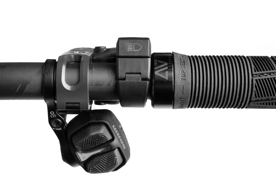 Nahaufnahme eines ACID E-Bike-Frontlichts PRO-E 200 High Beam X-Connect, strukturierter schwarzer Griff, Gangschaltung und X-Connect-Bedientasten an einem Fahrradlenker, alles vor einem weißen Hintergrund.