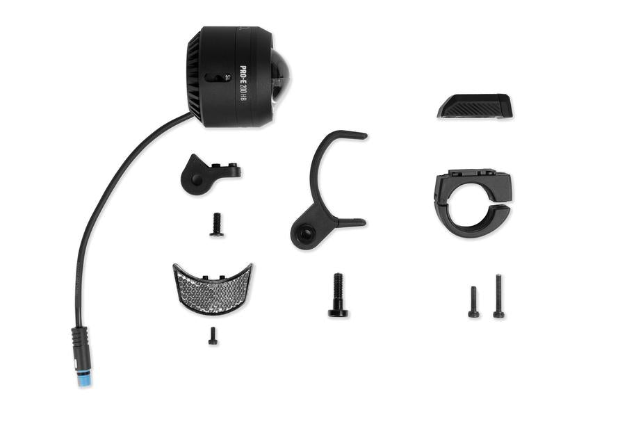 Das ACID E-Bike Frontlicht PRO-E 200 High Beam X-Connect schwarz ist im zerlegten Zustand abgebildet, wobei die Halterung, die Schrauben, zwei gebogene Aufsätze, eine reflektierende Platte und eine kleine Abdeckung ordentlich auf einem weißen Hintergrund angeordnet sind.