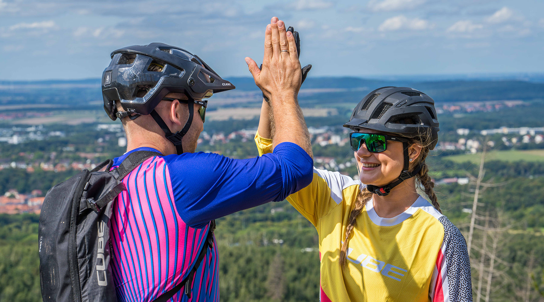 Zwei Radfahrer mit Helmen und Sonnenbrillen auf CUBE-Fahrrädern geben sich lächelnd ein High-Five, mit einer malerischen Aussicht auf Felder, Bäume und die ferne Stadt Göttingen in Niedersachsen im Hintergrund.