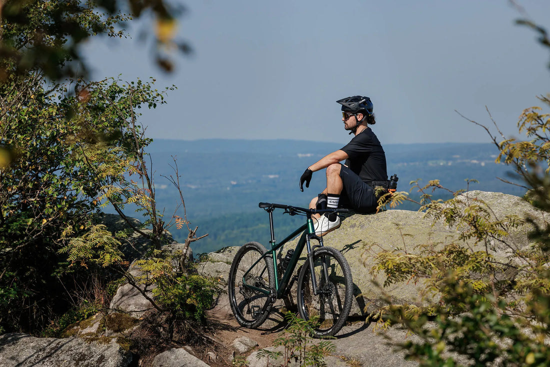 Mountainbiker macht Pause auf einem Felsen mit Panoramablick, daneben ein CUBE Hardtail-Mountainbike