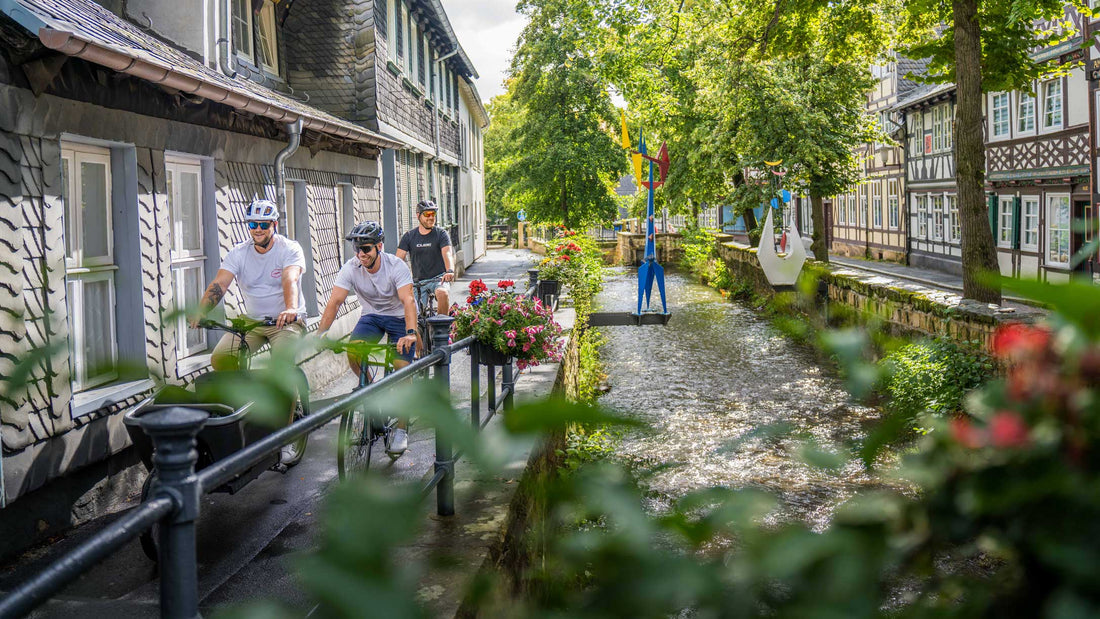 Drei Radfahrer fahren entlang eines Bachlaufs zwischen Fachwerkhäusern in einer Altstadt