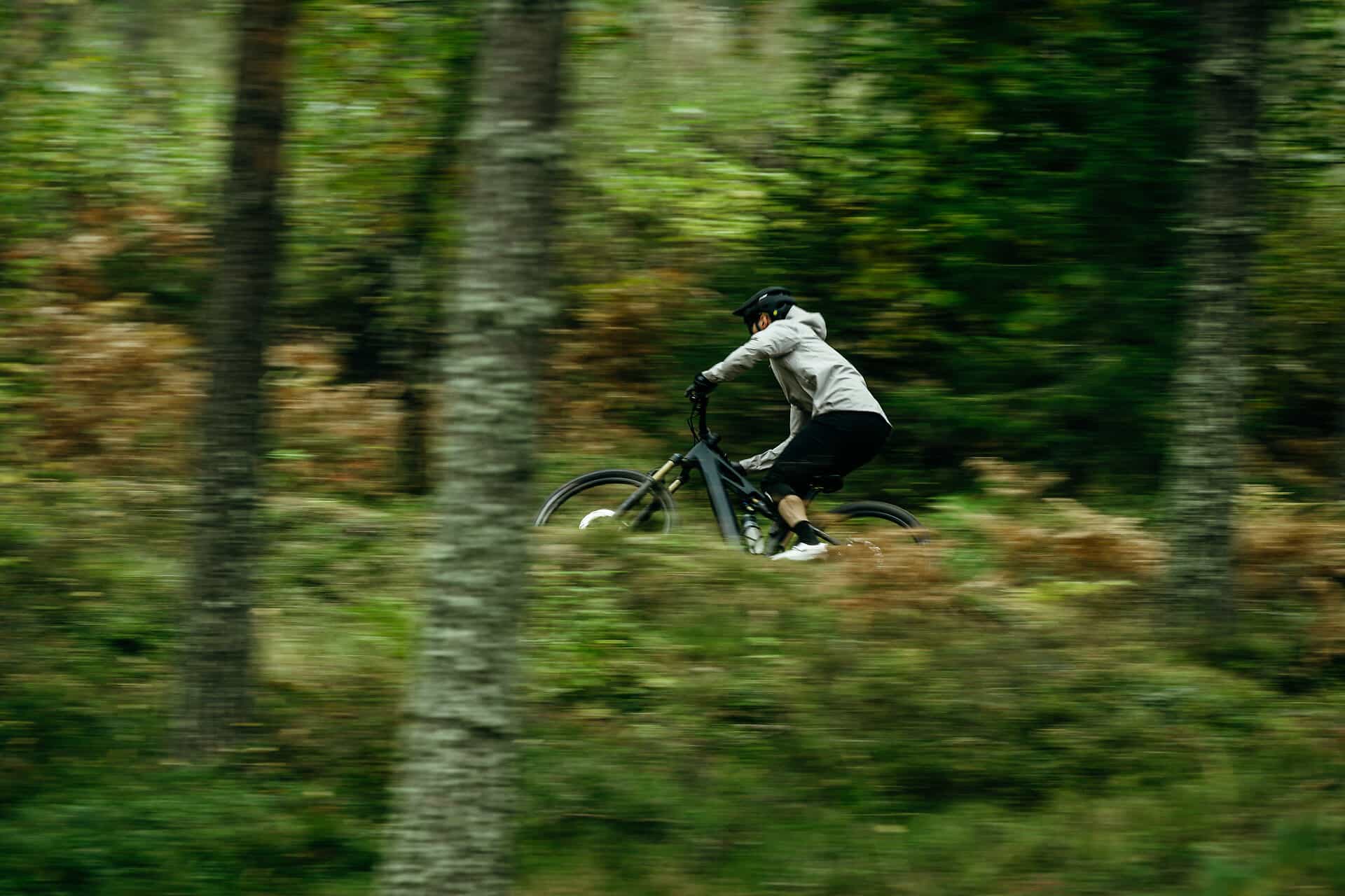 Eine Person, die einen Helm und eine Kapuze trägt, fährt mit einem Mountainbike schnell durch einen üppigen, grünen Wald, wobei die Bäume und das Laub verschwommen sind, was auf Bewegung hindeutet.