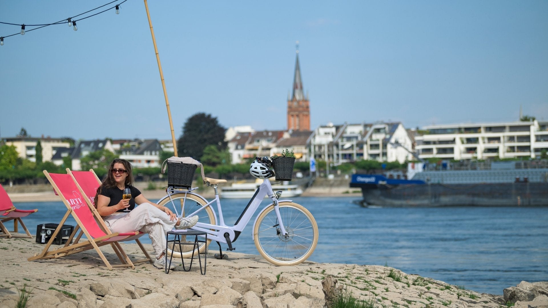 Frau sitz in einem klappstuhl und neben ihr steht ein fahrrad. sie ist im hafen und im hintergrund ist everschwommen ein schiff zu erkennen das in einem Kanal fährt.