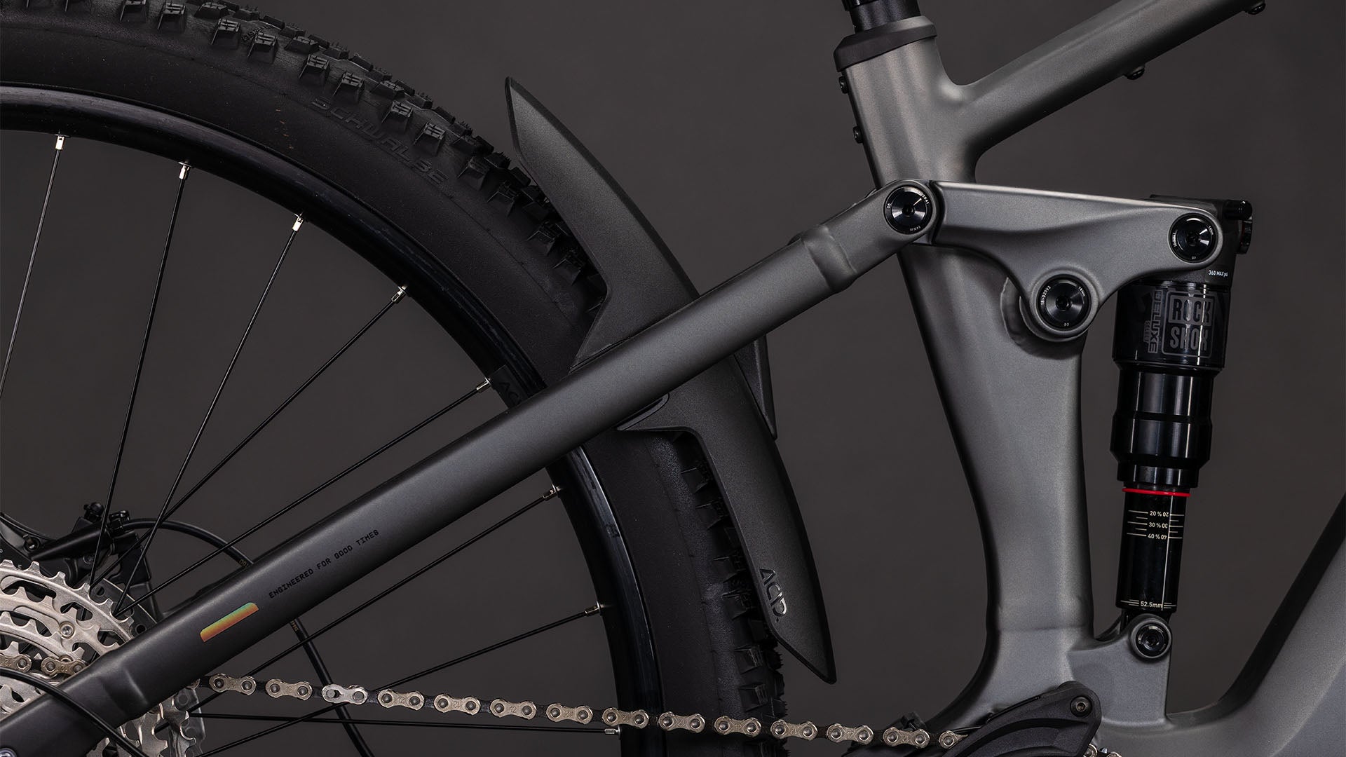 Nahaufnahme der Hinterradfederung und des Shimano XT-Antriebs des CUBE Stereo Hybrid ONE22 Race 800 slabgrey'n'chrome 2026 mit Hinterrad, Stoßdämpfer, Kette und mattschwarzem Schutzblech vor dunklem Hintergrund.