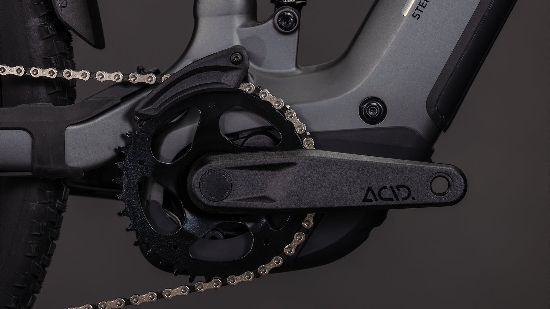 Nahaufnahme der Cube Stereo Hybrid ONE22 Race 800 slabgrey'n'chrome 2026 Kurbelgarnitur und Kettenblatt, mit einem ACID-gekennzeichneten Kurbelarm und Shimano 12-fach XT Schaltung von CUBE vor dunklem Hintergrund.