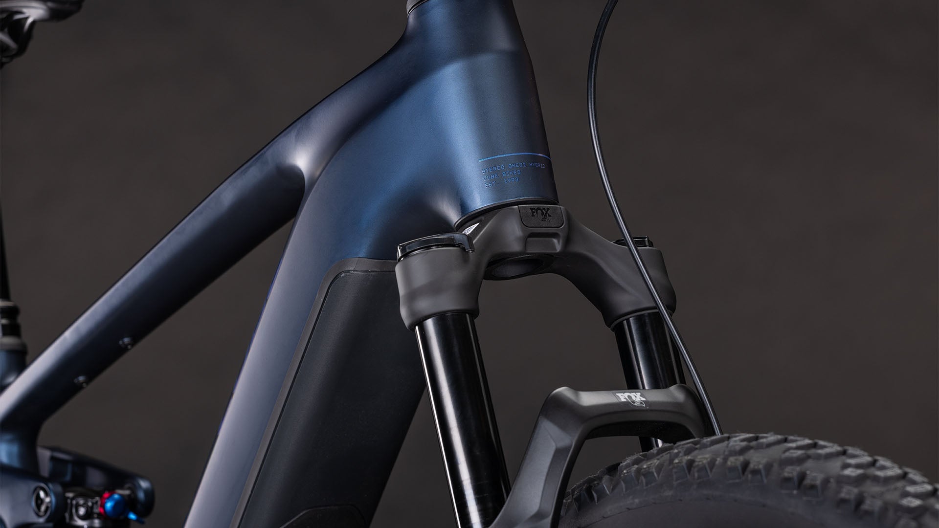 Nahaufnahme des mattblauen Rahmens und der Federgabel des CUBE Stereo Hybrid ONE22 SLX 800 indigoblue'n'blue 2026 mit Bosch CX-Motor und Shimano XT 12-Gang-Schaltung, dargestellt mit einem schwarzen Reifen vor dunklem Hintergrund.