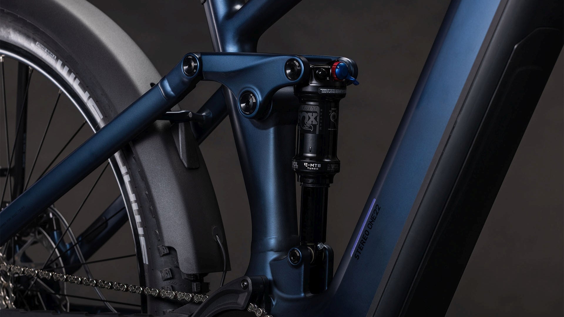 Nahaufnahme des CUBE Stereo Hybrid ONE22 SLX 800 FE indigoblue'n'blue 2026, die den mattblauen Rahmen, die Hinterradaufhängung mit Dämpfer und Gestänge, die sichtbare Kette, den Reifen, den Bosch CX Motor und die Details des Antriebs zeigt.