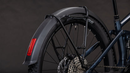 Nahaufnahme eines CUBE Stereo Hybrid ONE22 SLX 800 FE Hinterrads, die den blauen Metallrahmen, den Schwalbe-Reifen, das schwarze Schutzblech mit rotem Reflektor und einen Teil der Schaltung vor einem dunklen Hintergrund zeigt.