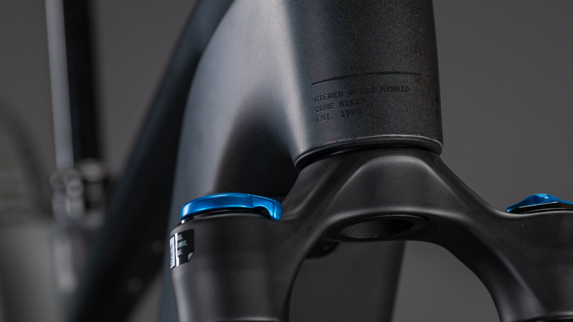 Nahaufnahme des CUBE Stereo Hybrid ONE22 SLT 800 prizmblack'n'black 2026 Rahmen und Gabel in mattem Schwarz, mit blauen Einstellknöpfen und einem dezent eingravierten Etikett, vor einem verschwommenen Hintergrund.
