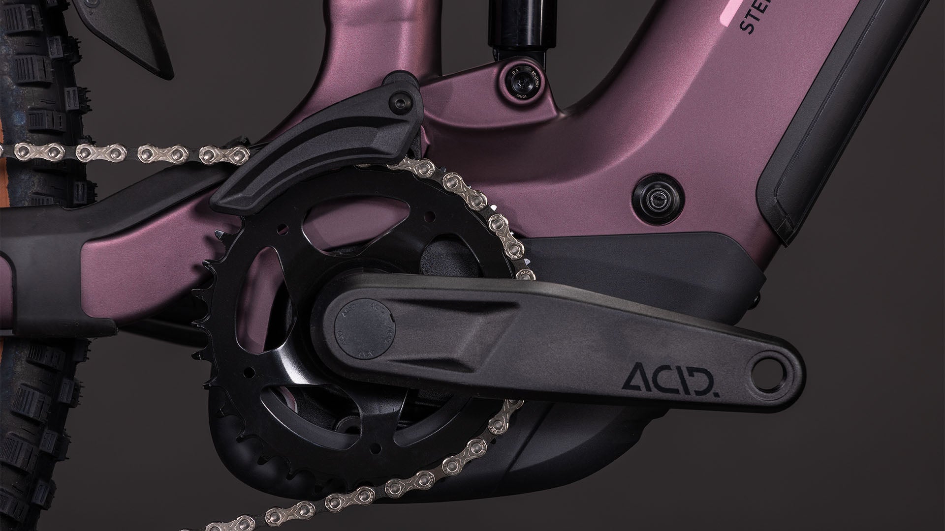 Nahaufnahme der Kurbelgarnitur und des Kettenblatts an einem mattlila CUBE Stereo Hybrid ONE44 Pro 800 Rahmen, Pedalarm mit der Aufschrift ACID, mit detaillierten Kettengliedern und einem dunklen Hintergrund, der die Shimano XT Schaltung hervorhebt.