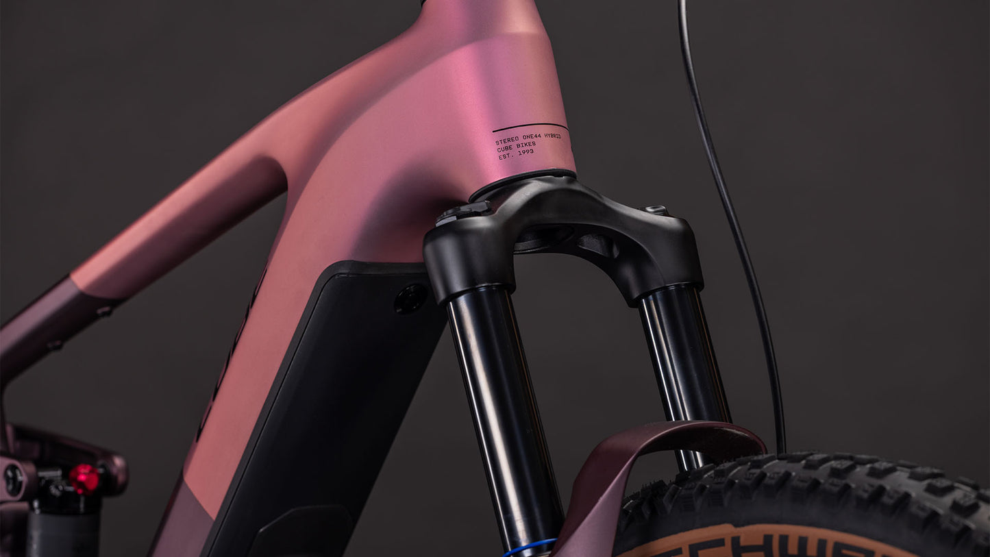 Nahaufnahme der vorderen Federgabel und des Rahmens eines CUBE Stereo Hybrid ONE44 Pro 800 shiftblush'n'raisin 2026 Mountainbikes, mit einem Teil des Vorderreifens vor einem dunklen Hintergrund.