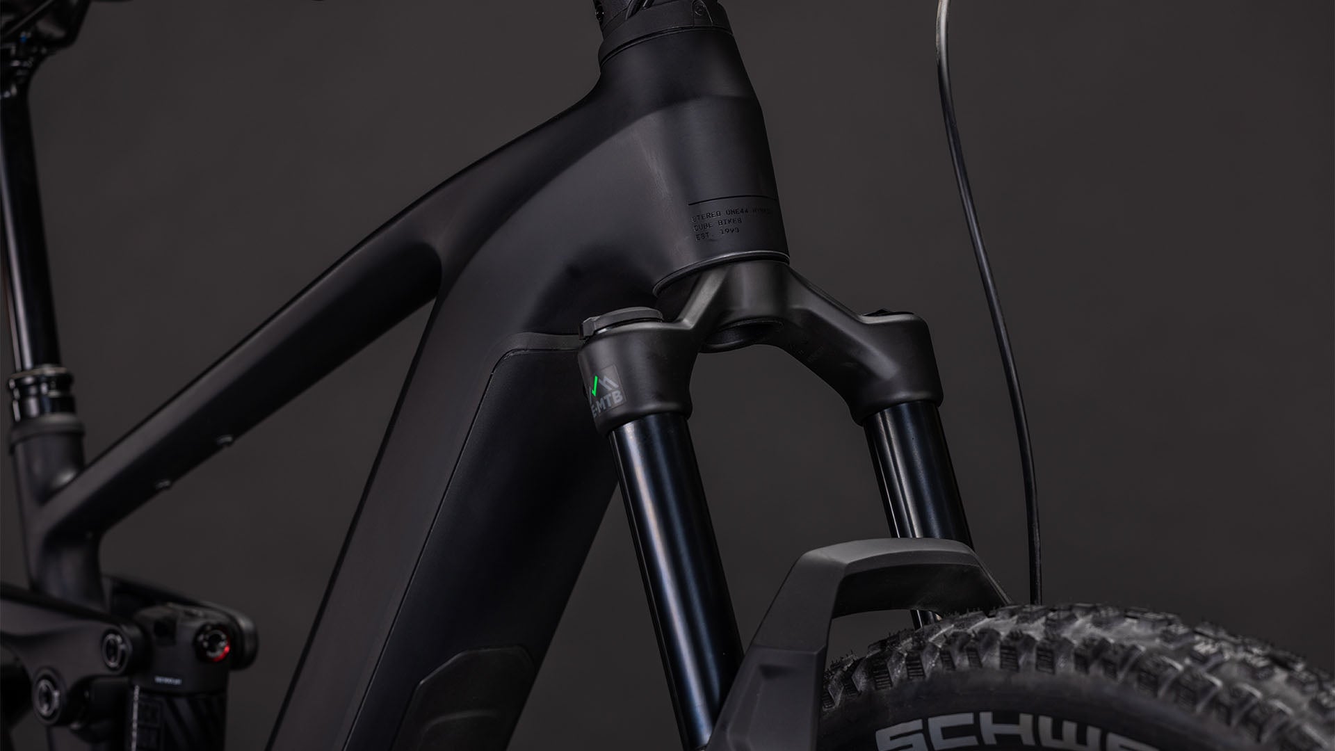 Nahaufnahme der vorderen Federgabel des CUBE Stereo Hybrid ONE44 EX 800 blackline 2026, des robusten Reifens mit sichtbarem Branding und eines Teils des klobigen Rahmens, alles vor einem dunklen Hintergrund, der das Fully-MTB-Design betont.
