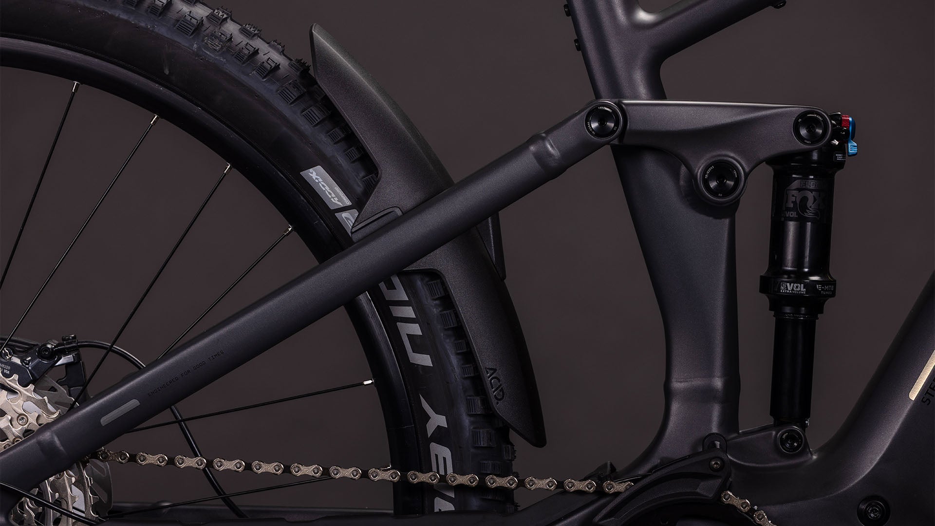 Nahaufnahme der Hinterradaufhängung, des Reifens, des Stoßdämpfers, der Kette und des hinteren Schutzblechs des CUBE Stereo Hybrid ONE44 EXC 800 irongrey'n'chrome 2026, ausgestattet mit einer Shimano XT Schaltung für reibungslose Schaltvorgänge vor einem dunklen Hintergrund.