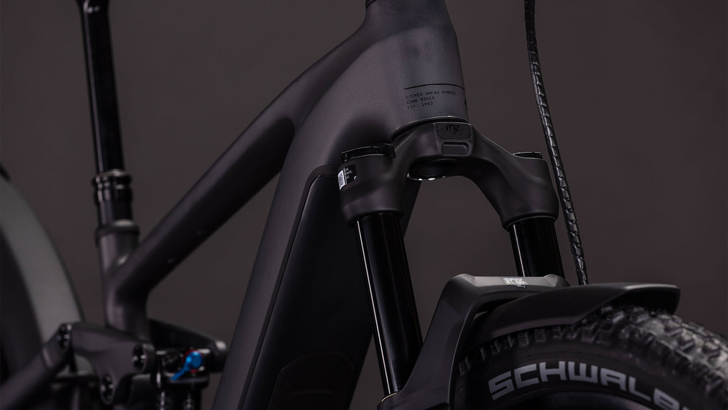Nahaufnahme eines matten, irongrünen und verchromten CUBE Stereo Hybrid ONE44 EXC 800 FE Mountain E-Bikes, das die vordere Federgabel, den Rahmen, die Schwalbe-Reifen und den Bosch CX-Motor vor einem dunklen Hintergrund zeigt.