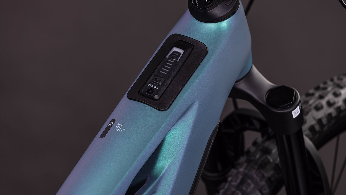 Nahaufnahme des Cube Stereo Hybrid ONE44 HPC Race 800 smaragdgrey'n'prism von CUBE, die das integrierte Bedienfeld und die Knöpfe am High Performance Carbon-Rahmen zeigt, sowie einen Teil der Federgabel und des Noppenreifens.
