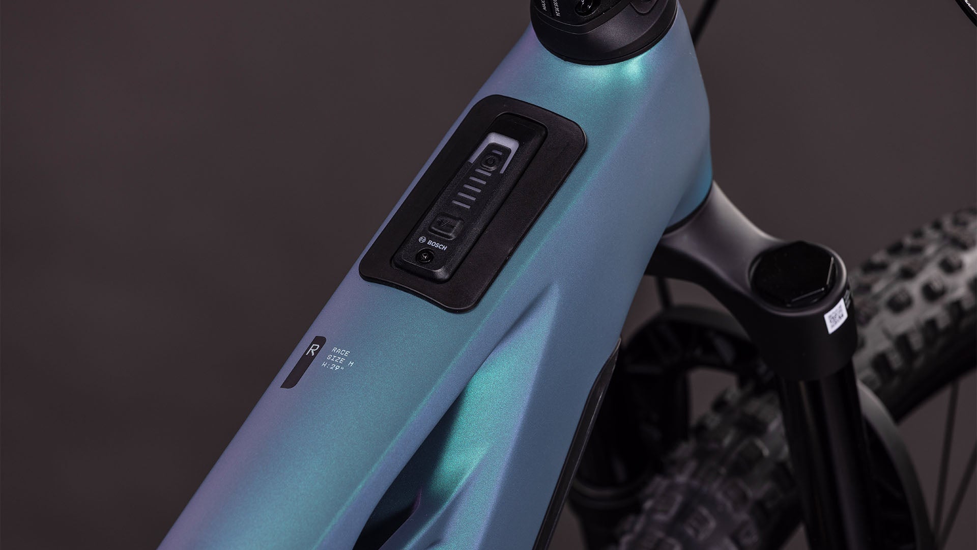 Nahaufnahme des Cube Stereo Hybrid ONE44 HPC Race 800 smaragdgrey'n'prism von CUBE, die das integrierte Bedienfeld und die Knöpfe am High Performance Carbon-Rahmen zeigt, sowie einen Teil der Federgabel und des Noppenreifens.