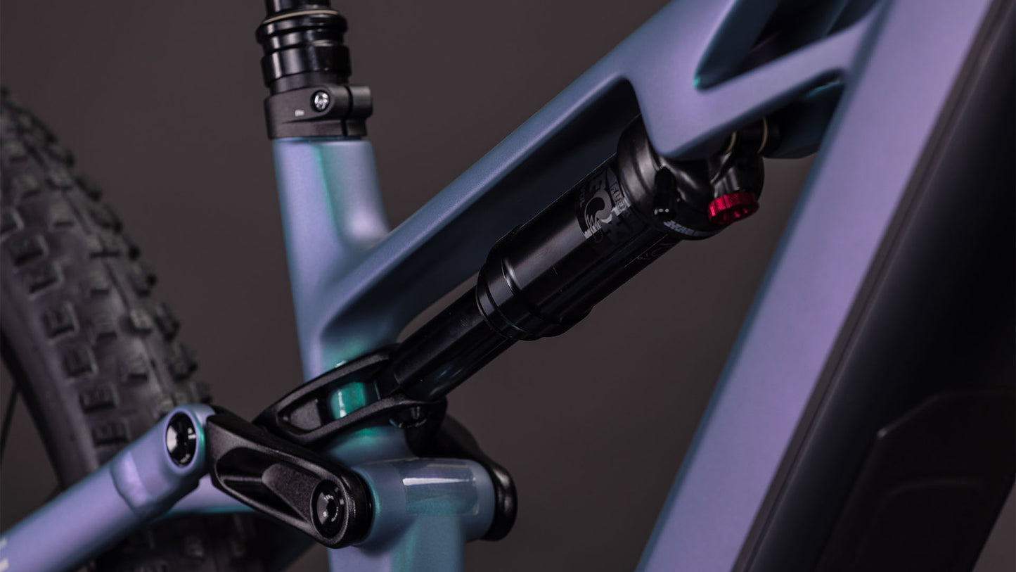 Nahaufnahme der Hinterradaufhängung des CUBE Stereo Hybrid ONE44 HPC Race 800. Hervorzuheben sind der Stoßdämpfer und das Gestänge des Hochleistungs-Carbonrahmens sowie ein Teil des Hinterradreifens vor einem dunklen Hintergrund.