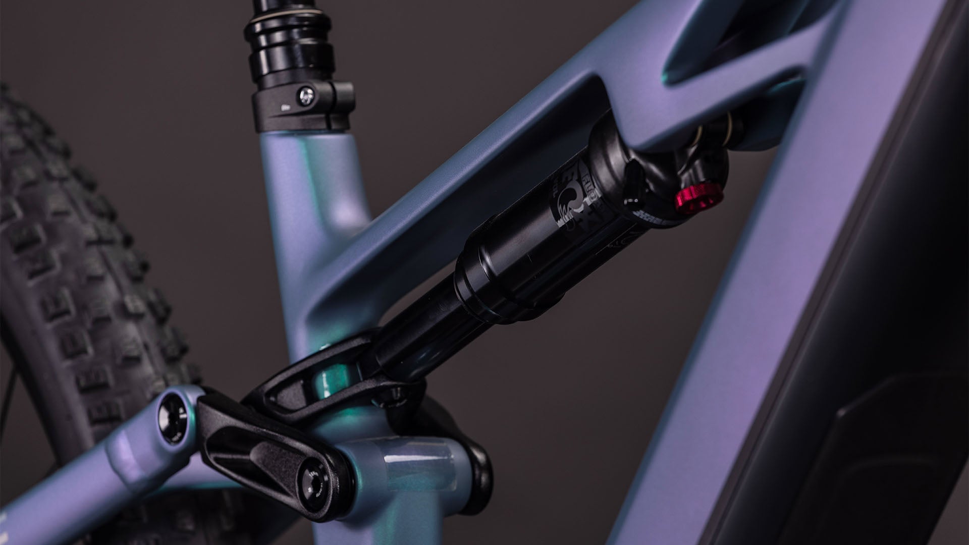 Nahaufnahme der Hinterradaufhängung des CUBE Stereo Hybrid ONE44 HPC Race 800. Hervorzuheben sind der Stoßdämpfer und das Gestänge des Hochleistungs-Carbonrahmens sowie ein Teil des Hinterradreifens vor einem dunklen Hintergrund.