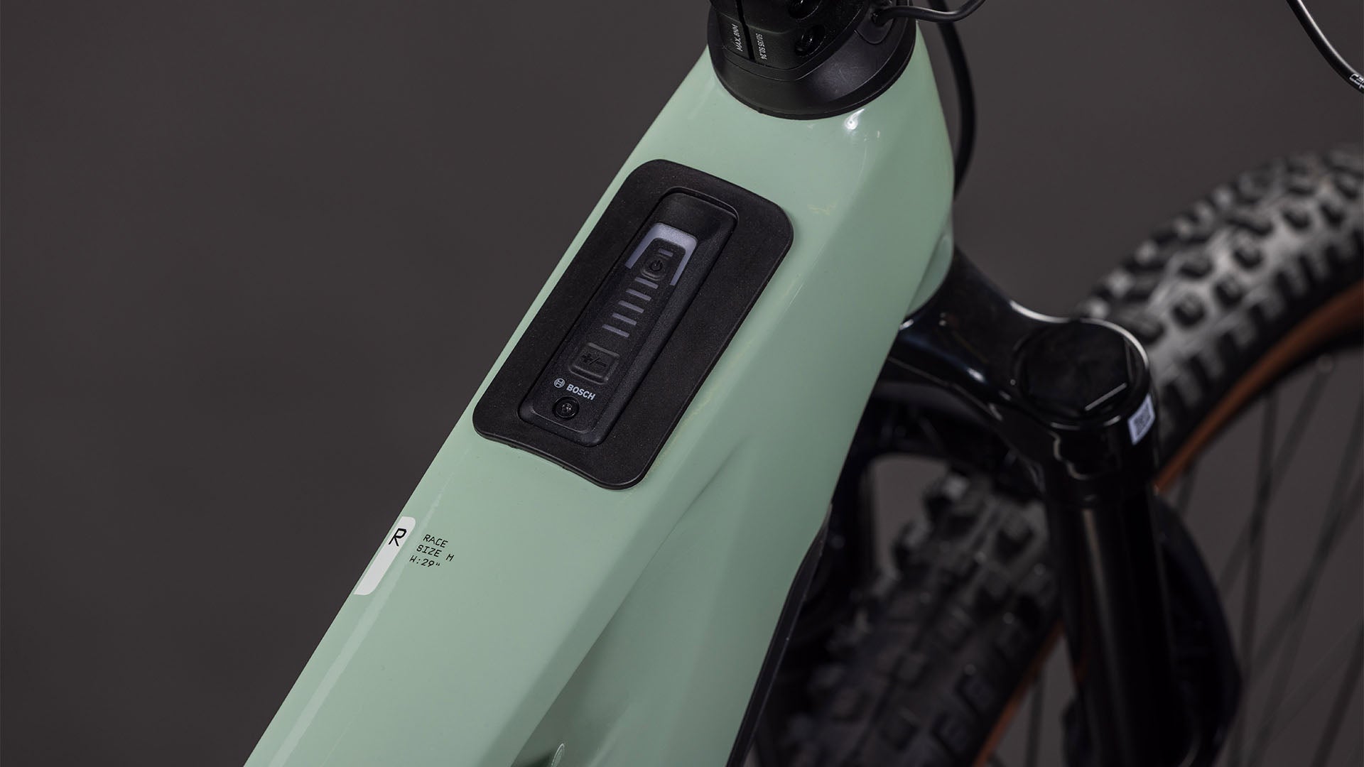 Nahaufnahme des CUBE Stereo Hybrid ONE44 HPC Race 800 driedherbs'n'black 2026-Rahmens mit integrierter Bosch-Akku-Ladeanzeige und Powerknopf. Vorderrad und Lenker weisen auf das leistungsstarke Carbon-Design hin.