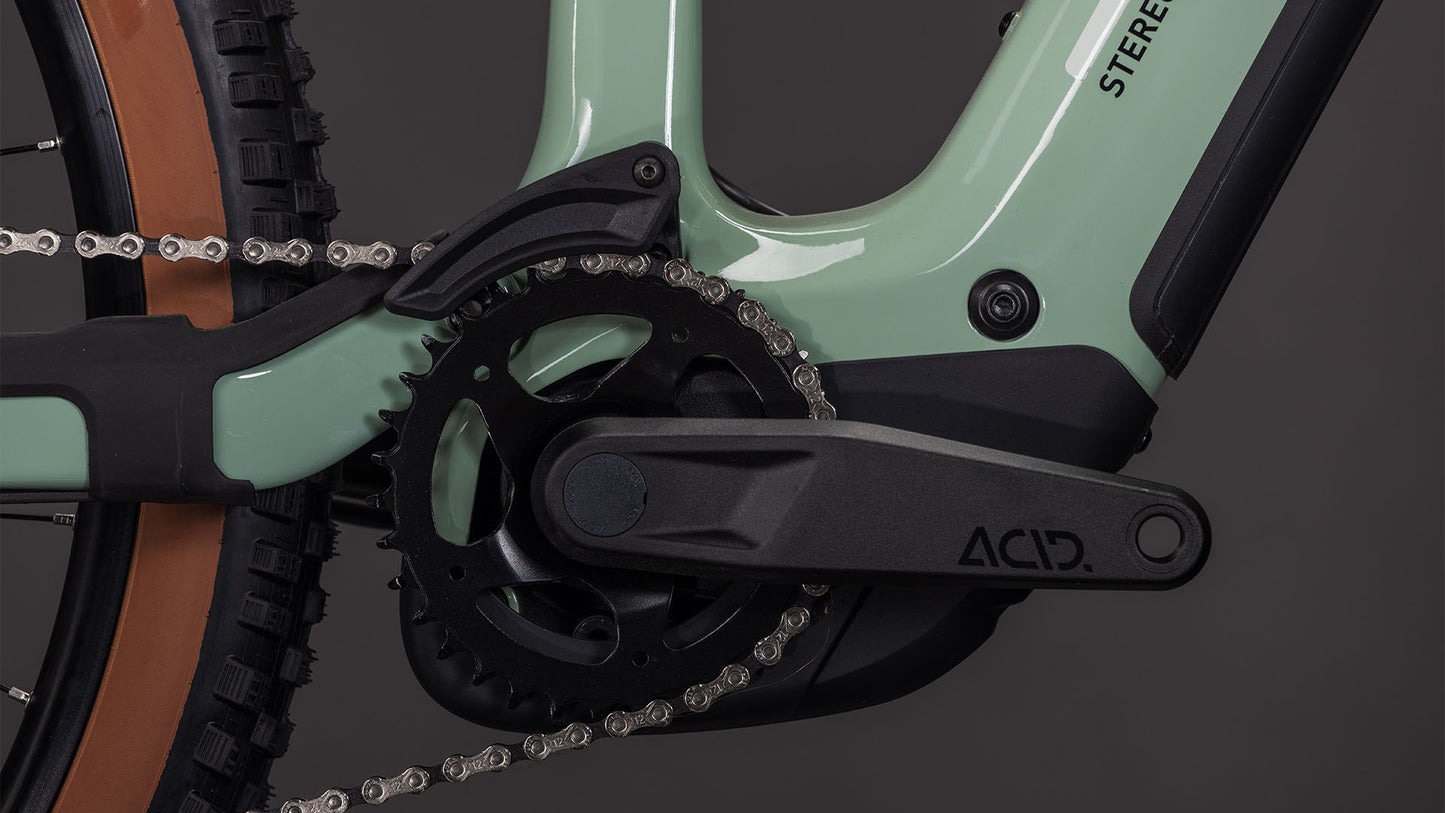 Nahaufnahme der High-Performance-Carbon-Kurbelgarnitur und des schwarzen ACID-Kurbelarms des CUBE Stereo Hybrid ONE44 HPC Race 800 driedherbs'n'black 2026, die am mintgrünen Rahmen befestigt sind, wobei ein Teil des Hinterrads und des Reifens sichtbar ist.