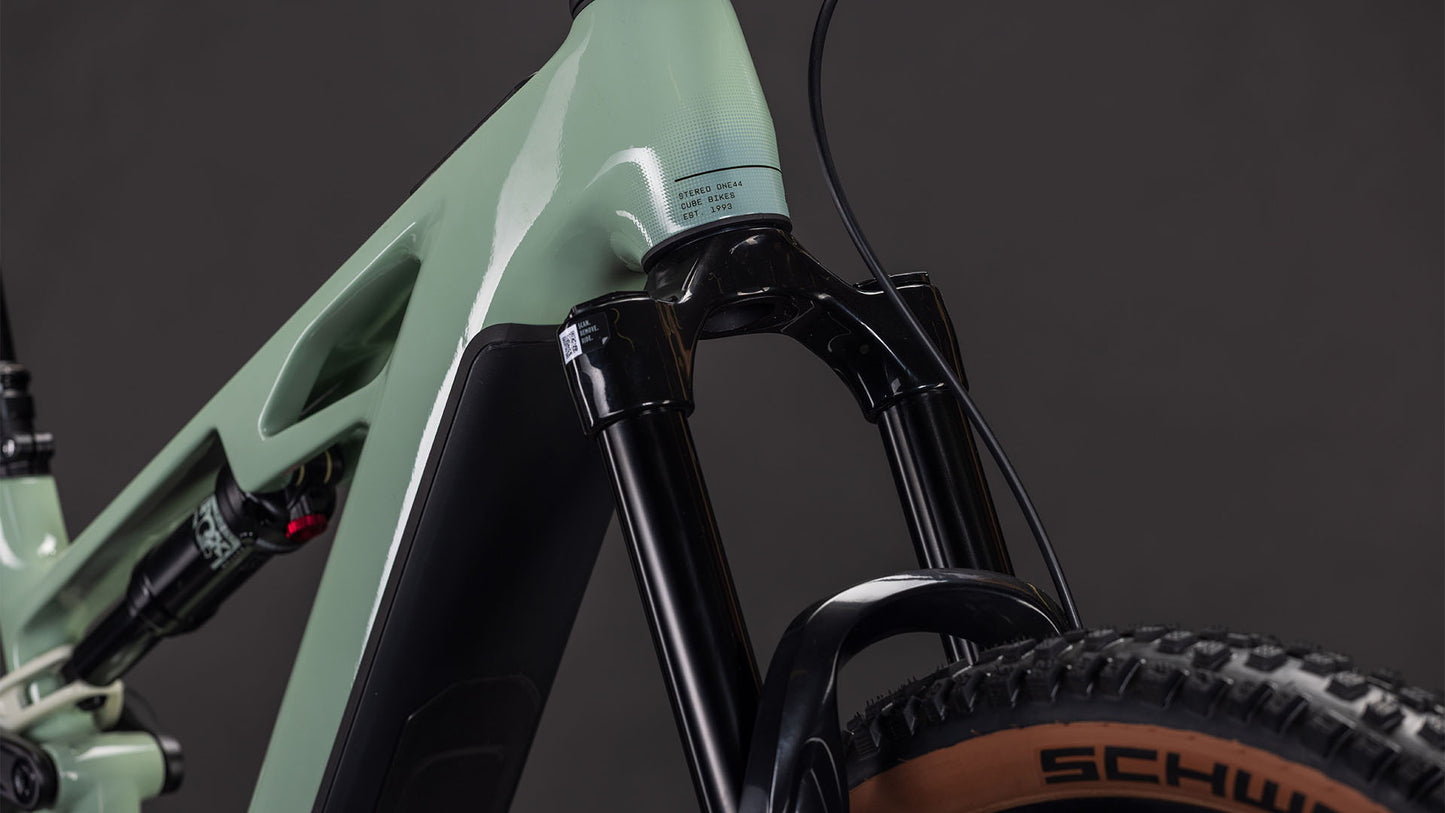Nahaufnahme eines CUBE Stereo Hybrid ONE44 HPC Race 800 driedherbs'n'black 2026 mit Federgabel und Reifen der Marke Schwalbe, mit einem matt-hellgrünen High Performance Carbon-Rahmen und schwarzen Komponenten vor einem dunklen Hintergrund.