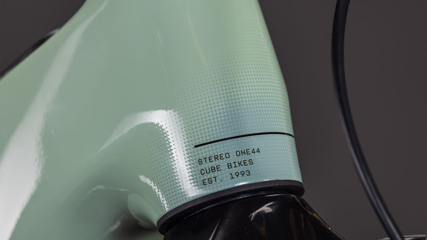 Nahaufnahme des CUBE Stereo Hybrid ONE44 HPC Race 800 driedherbs'n'black 2026 Rahmens mit gepunkteter Textur und "STEREO ONE44 CUBE BIKES EST. 1993" in der Nähe des Steuersatzes; daneben verläuft ein schwarzes Kabel.