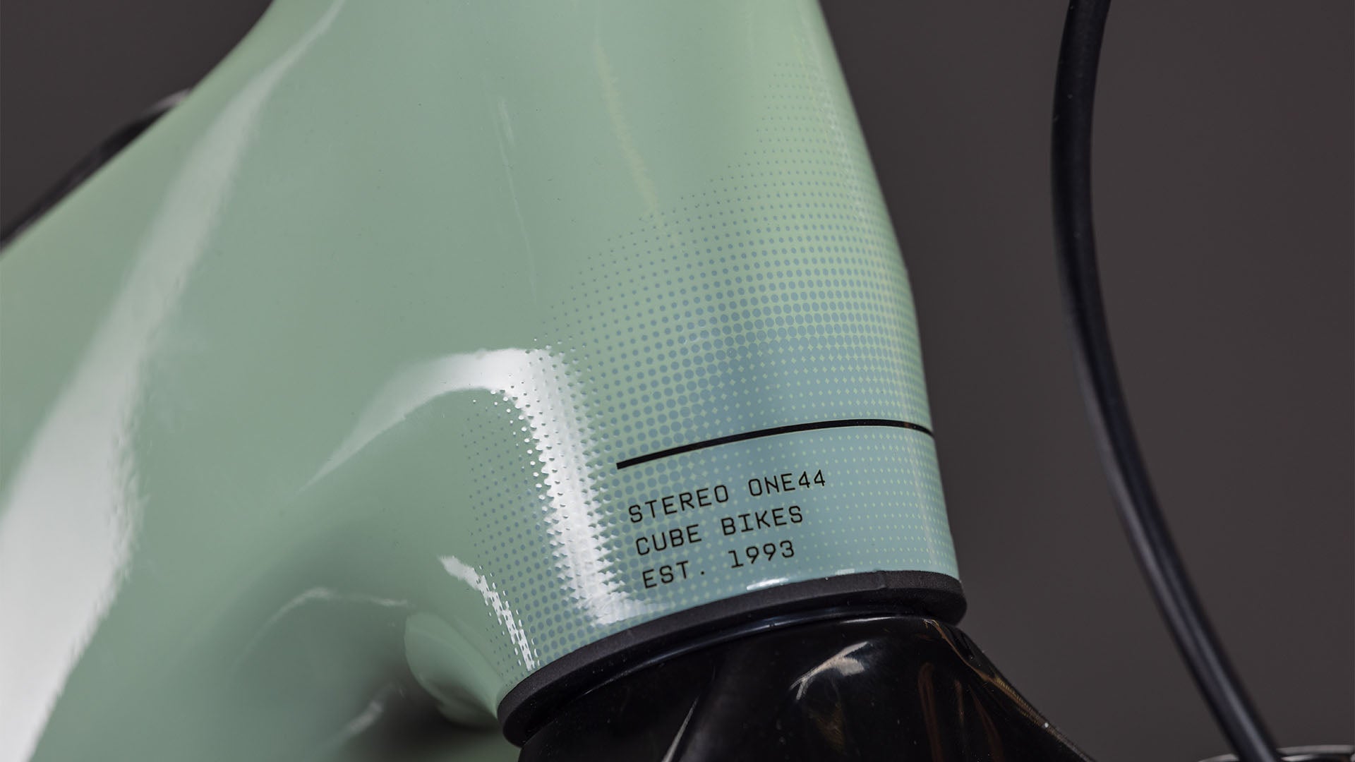 Nahaufnahme des CUBE Stereo Hybrid ONE44 HPC Race 800 driedherbs'n'black 2026 Rahmens mit gepunkteter Textur und "STEREO ONE44 CUBE BIKES EST. 1993" in der Nähe des Steuersatzes; daneben verläuft ein schwarzes Kabel.